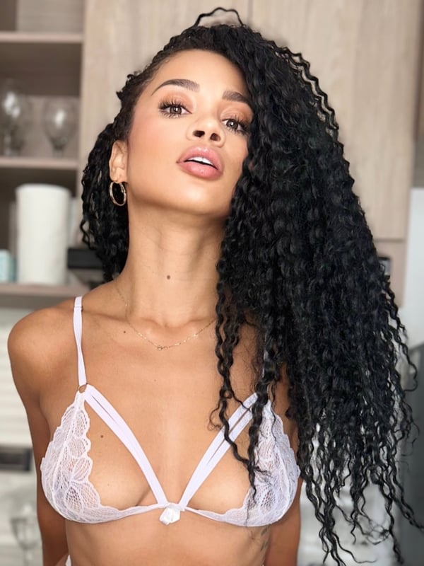 A woman with curly hair smiling and posing in a kitchen area with cabinets and glassware.