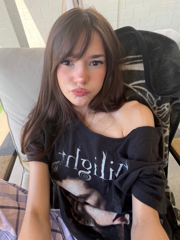 A young woman with long brown hair and bangs taking a mirror selfie while wearing a pink jacket and holding a decorated phone case.