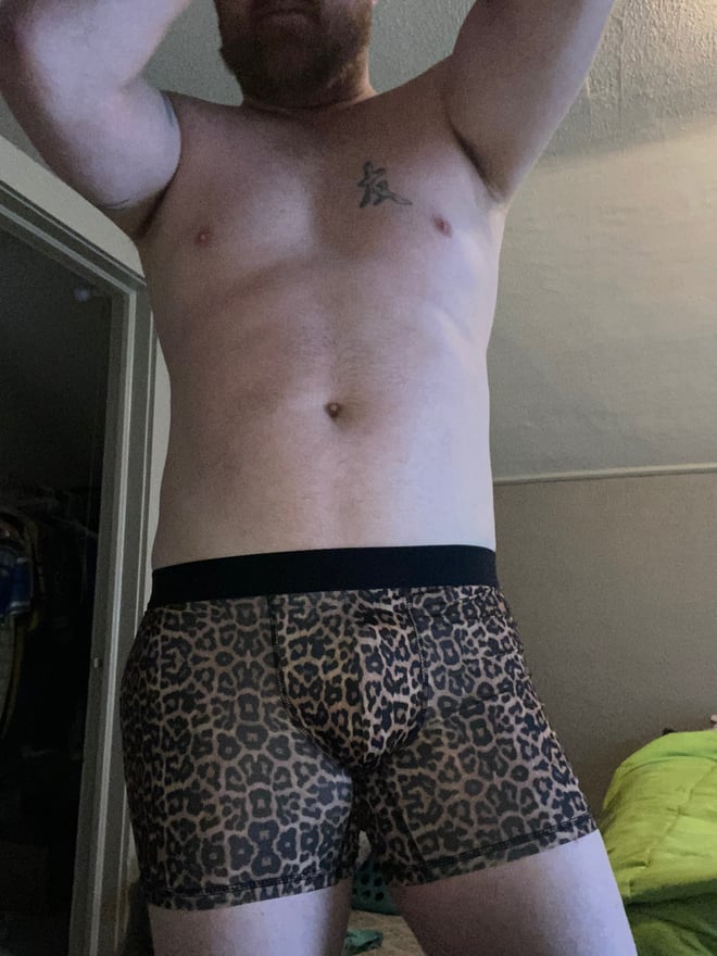 A muscular man standing shirtless in leopard print underwear, posing with arms raised in a bedroom setting.