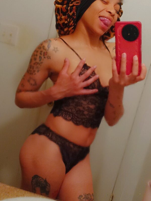 Woman with leopard print hair wrap taking a selfie in lingerie, showing tattoos on her arms and legs in a bathroom mirror.