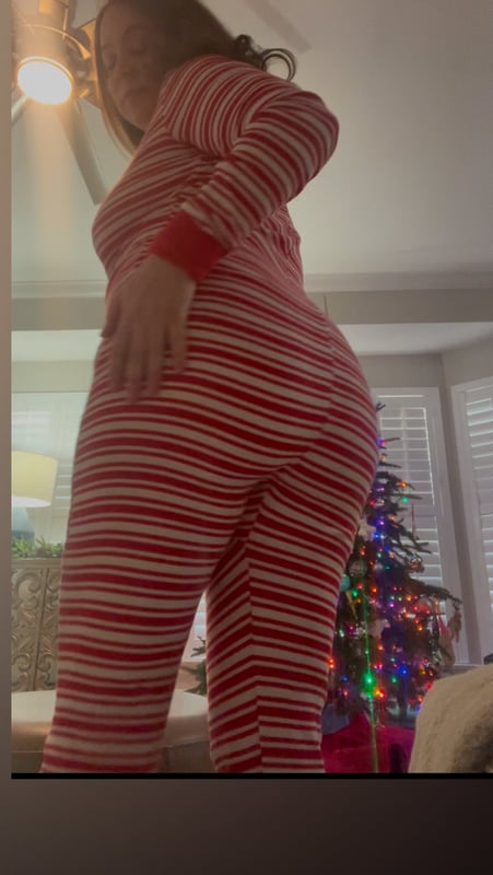 A woman posing in red and white striped pajamas in front of a Christmas tree and mirror in a cozy room.