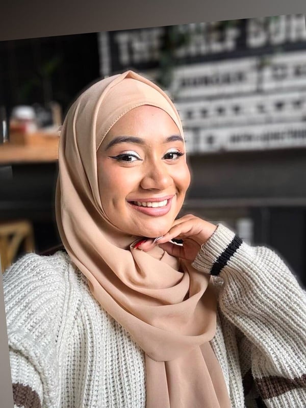 Smiling woman wearing a beige hijab and white sweater, posing with her hand on her cheek in a cozy indoor setting with plants.