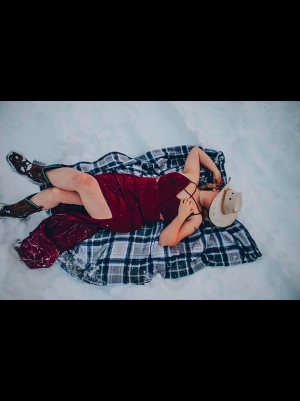 A woman with long blonde hair lies on a plaid blanket outdoors, wearing a red dress, black boots, and a cowboy hat.