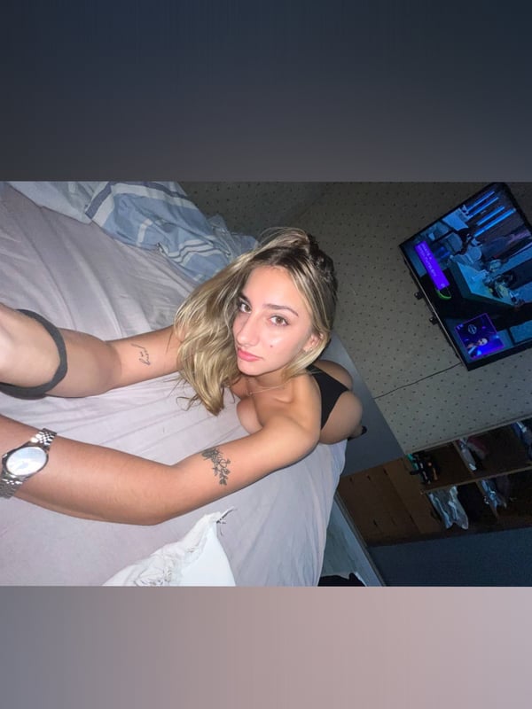 Young woman with blonde hair lounging on a bed in a dimly lit room with a TV in the background.