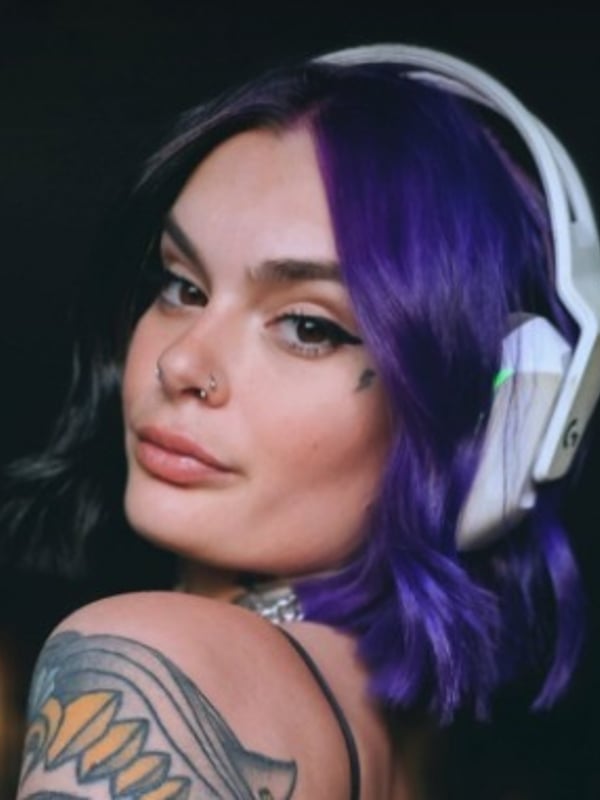A close-up of a woman with purple and black dyed hair, headphones, facial piercings, and tattoos, posing with a sultry expression against a dark background.