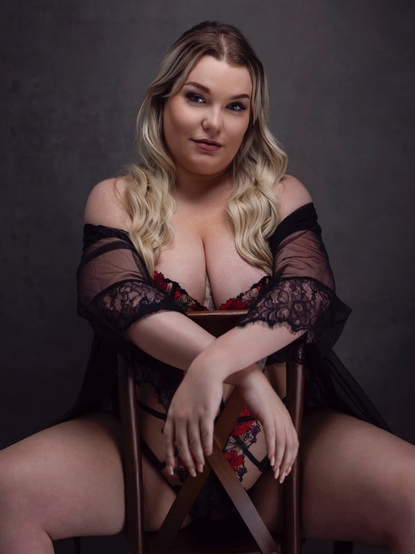 A woman with blonde hair sitting with legs crossed, dressed in black lace lingerie with a sheer robe, against a dark studio background.
