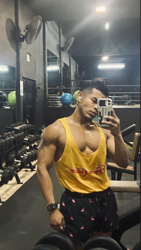 A muscular man in a yellow tank top and pink shorts posing in a gym with exercise equipment and mirrors in the background.