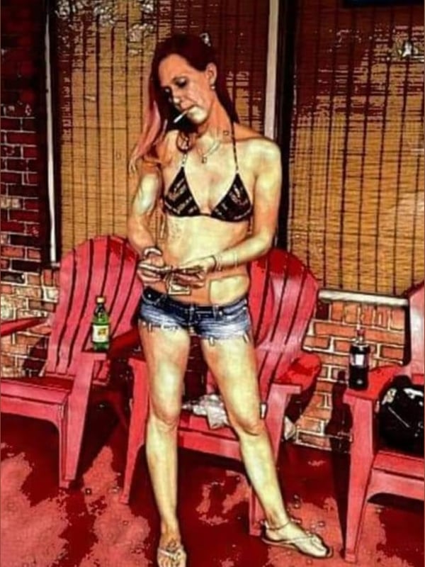 A woman with reddish hair wearing a black bikini top, short denim shorts, and flip-flops, posing confidently on a porch near a tattoo parlor sign with chairs and beer bottles nearby.