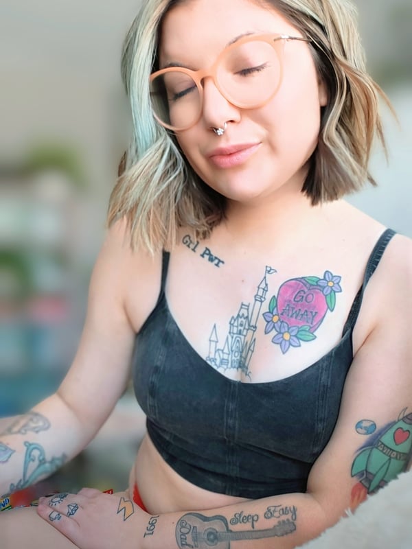 Young woman with dyed blonde and orange hair wearing orange-tinted glasses and a black top, sitting indoors with visible tattoos on her arms and nose piercing.