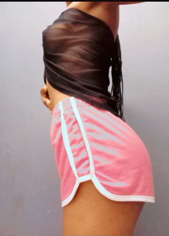 A woman posing sideways in a sheer top and pink shorts against a plain background.