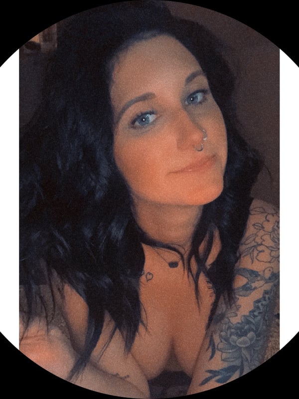 Close-up portrait of a woman with dark wavy hair, blue eyes, and tattoos on her shoulder, smiling softly.