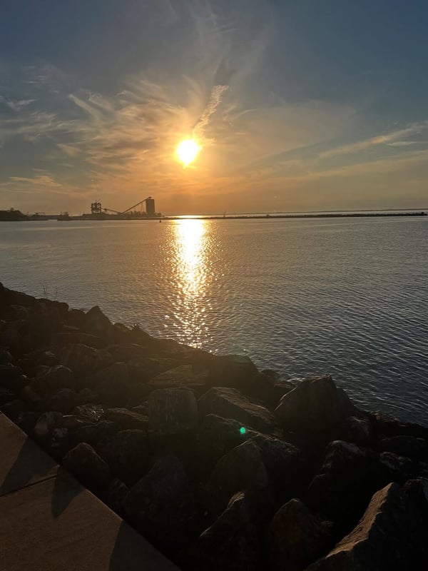 Scenic sunset over a body of water with industrial structures silhouetted against the horizon and rocky shoreline in the foreground.