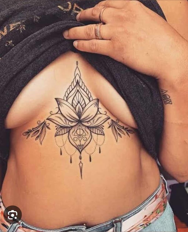 A woman posing in a loose gray top, revealing intricate floral tattoos on her chest and abdomen, with hands gently placed on her clothing.