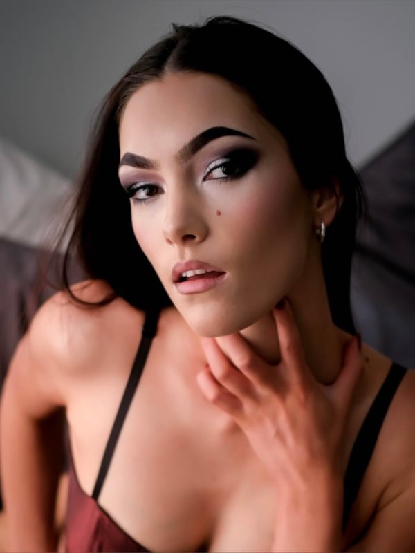 A woman with dark hair and dramatic makeup posing close-up with her hand near her face against a neutral background.
