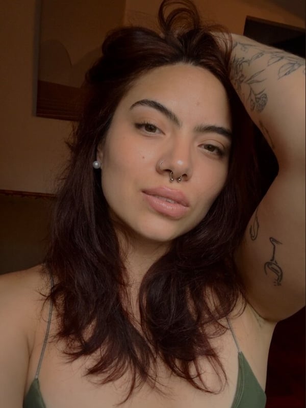 A woman with long brown hair taking a close-up selfie in a green top, arm raised showing tattoos, against a warm indoor background.