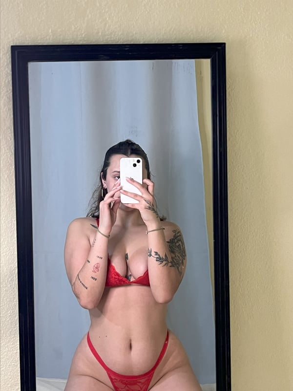 A young woman taking a mirror selfie in a red bikini swimsuit, standing in front of a black-framed mirror with tattoos on her arms.