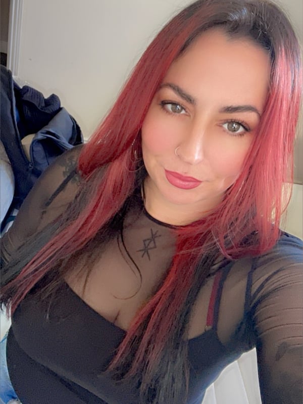 A woman with long red hair smiling confidently at the camera, wearing a sheer black top and jeans in a home interior.