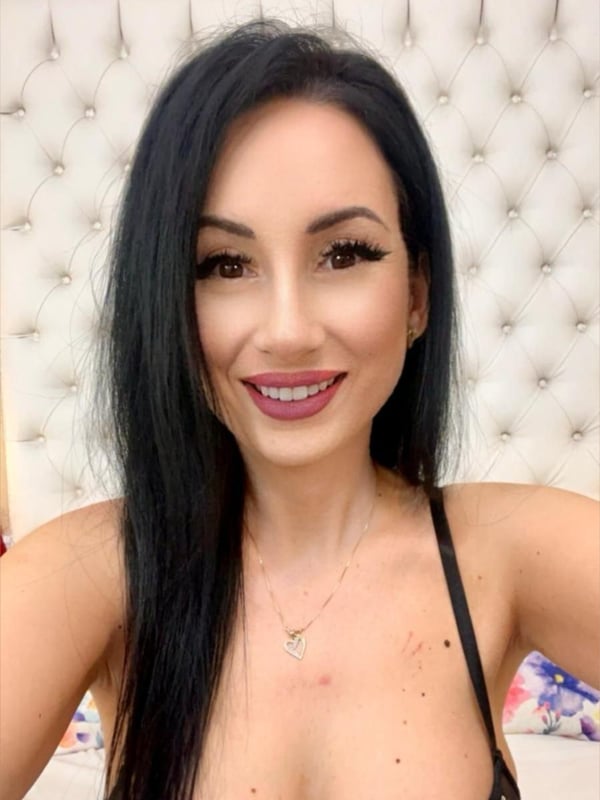 A woman with long dark hair smiling at the camera, wearing a low-cut top against a beige headboard background.