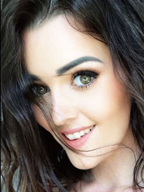 Close-up portrait of a smiling young woman with long wavy brunette hair and green eyes, wearing a delicate necklace.