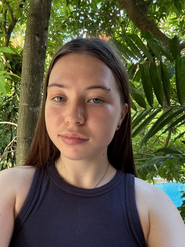 A close-up portrait of a young woman with long brown hair, freckles, and facial piercings, smiling slightly in front of green foliage.
