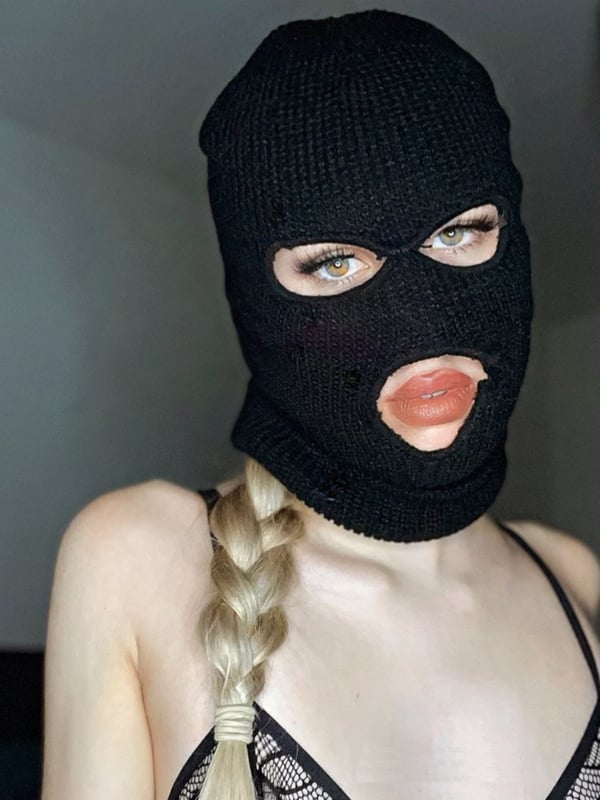 A person wearing a black balaclava mask and lace top, with blonde braided hair, posing confidently indoors.
