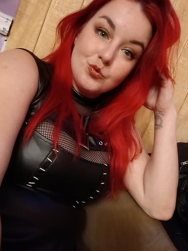 Close-up selfie of a woman with long red hair, green eyes, and a playful expression, dressed in a black mesh and leather top against a wooden background.