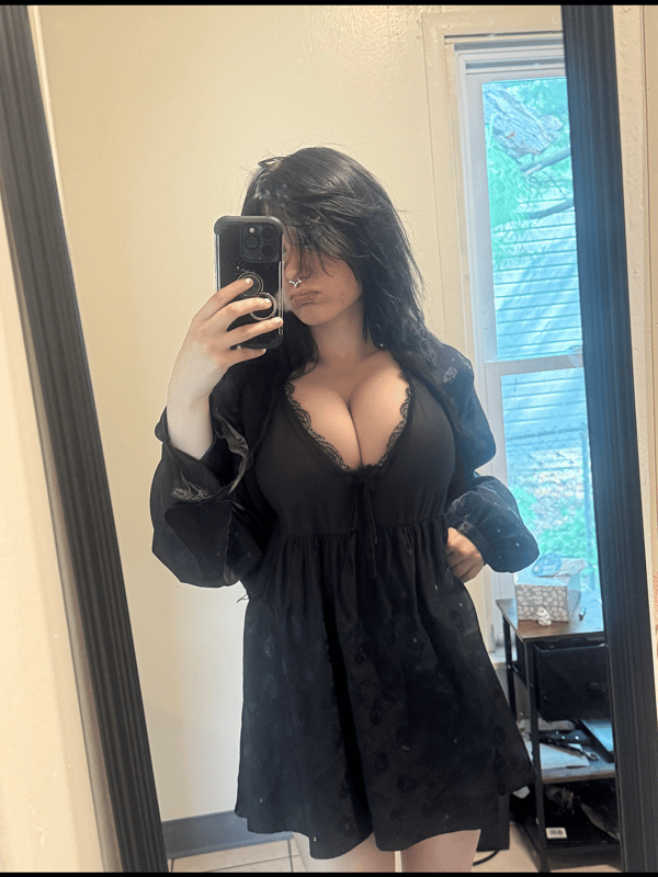 Young woman with long dark hair taking a mirror selfie in a low-cut black mini dress, standing indoors near a window.
