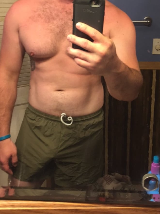 A muscular man taking a selfie in swim trunks, standing in a bathroom with blue walls and towels.