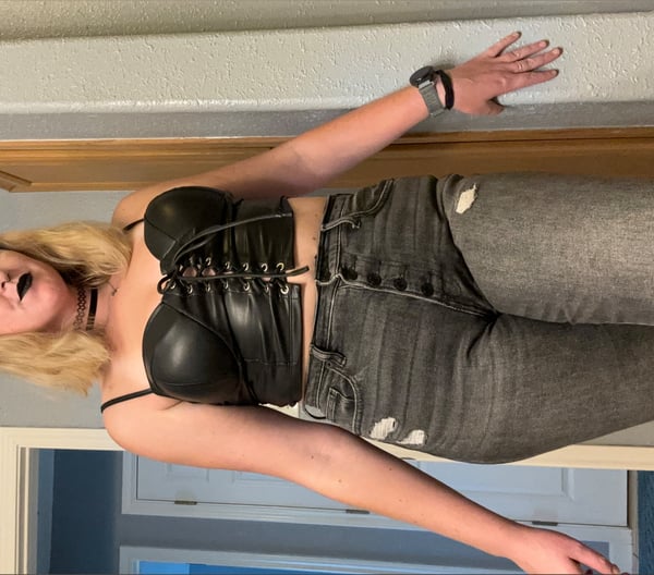 Young woman with blonde hair posing confidently indoors, wearing a black corset-style top and distressed jeans against a wooden surface.