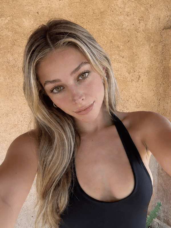 A close-up selfie of a young woman with long blonde hair wearing a black tank top, standing outdoors against a stone wall.
