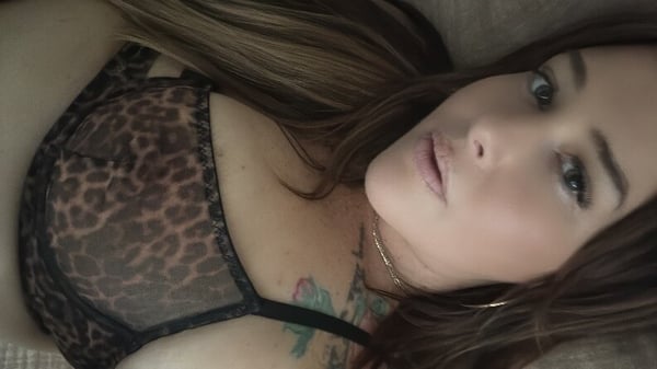 A woman with long hair reclining on a surface, wearing a patterned top and jewelry, with tattoos visible on her chest.