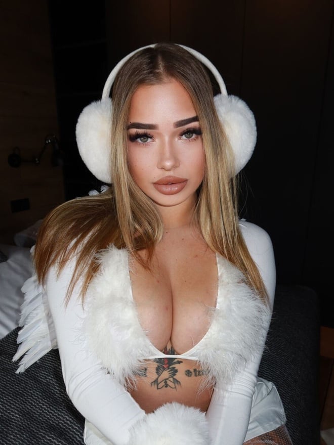 A woman with long blonde hair wearing fluffy earmuffs and a white fuzzy top, posing confidently in a bedroom setting.
