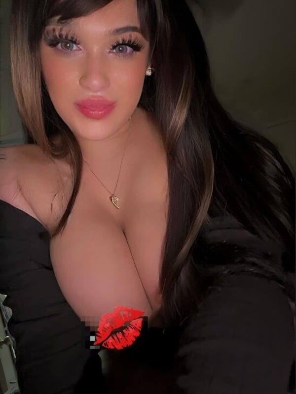 Woman with long dark hair and bold makeup posing confidently in a low-cut black top with a red lip detail.