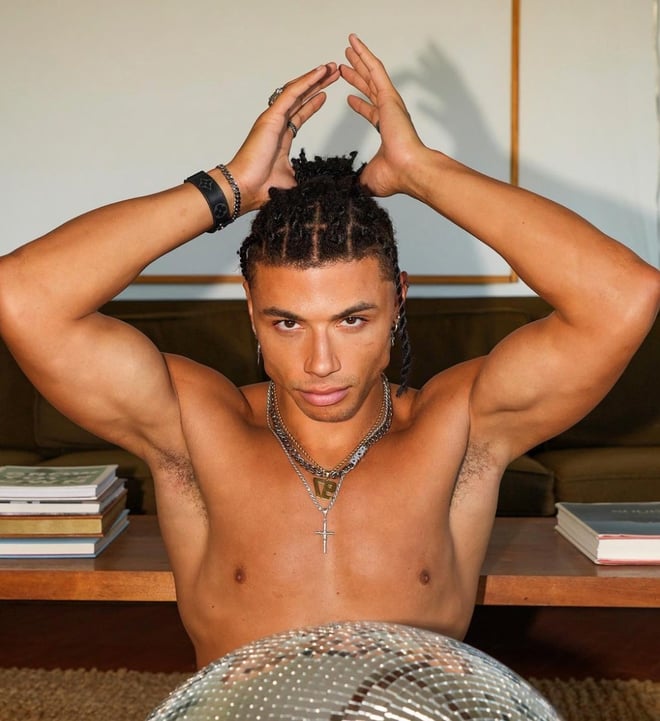 A muscular person with braided hair poses indoors, wearing jewelry and sitting behind a reflective silver surface.