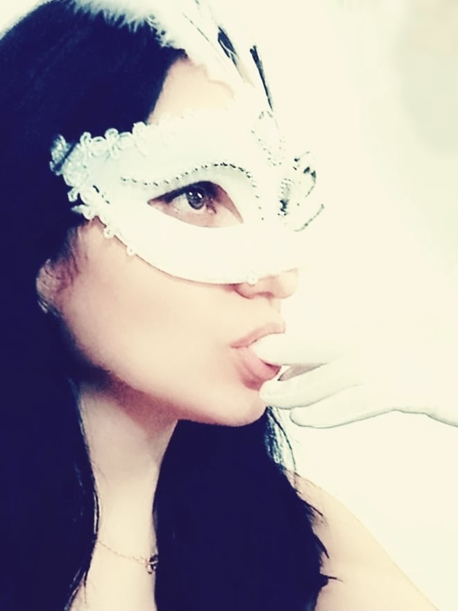 A woman wearing a white lace mask and feather accents, puckering her lips while holding a white object, with long dark hair.