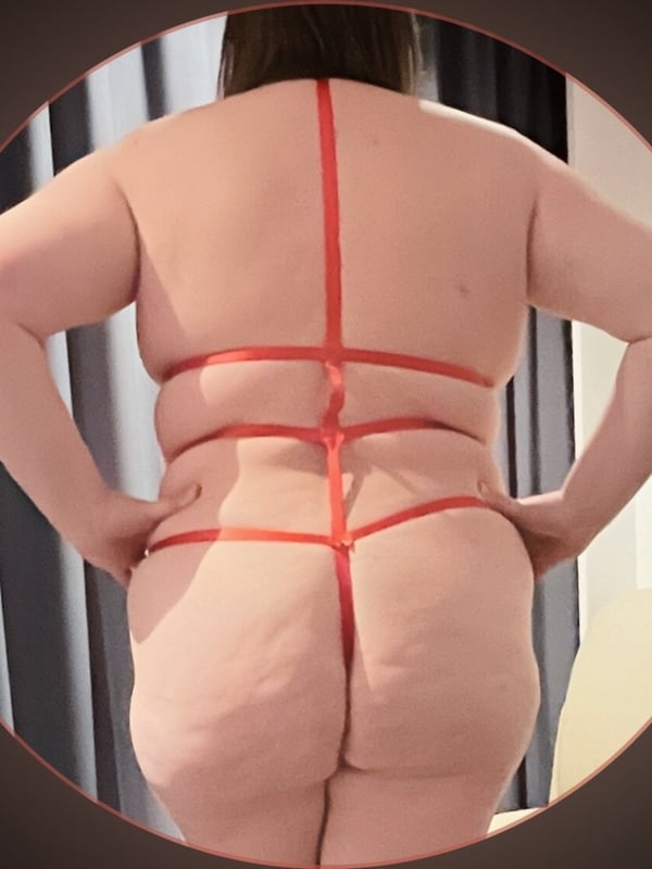 Woman standing with back view wearing red harness-style lingerie in a room.