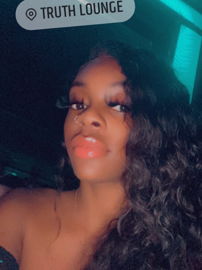 A smiling woman with long wavy hair posing confidently in a vibrant neon-lit lounge setting.