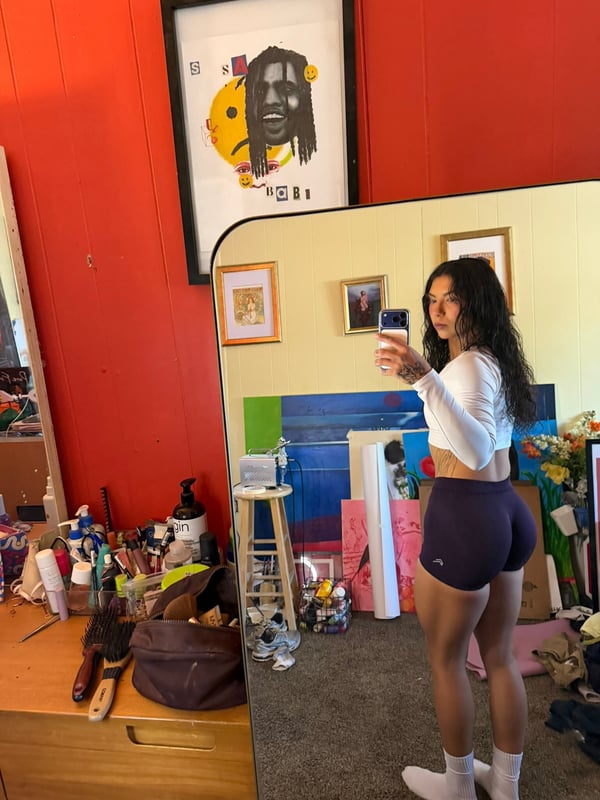 A woman taking a mirror selfie in a colorful room, wearing a white crop top and dark shorts.