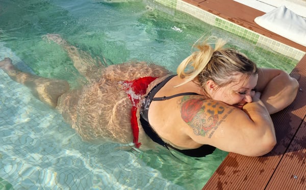 A woman with blonde hair reclining in a swimming pool, wearing a red bikini top and bottoms, with tattoos visible on her arm.