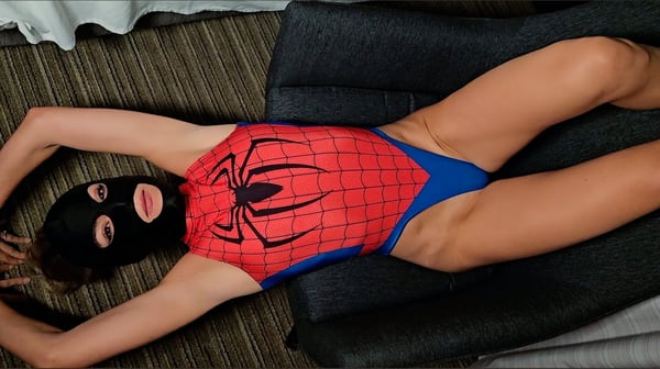 A person in a red and blue Spider-Man themed costume with a black mask, posing on a dark couch in a room with curtains.