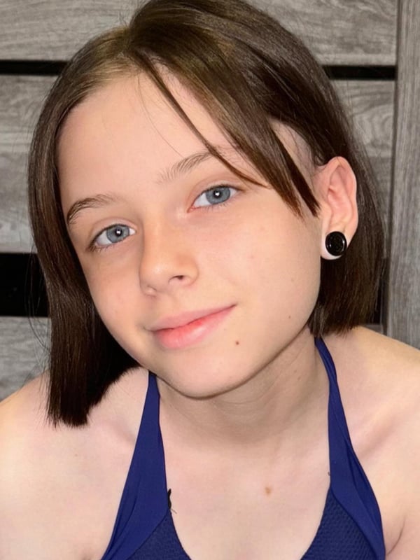 Close-up portrait of a young woman with short brown hair and piercings, smiling while wearing a blue top against a wooden background.