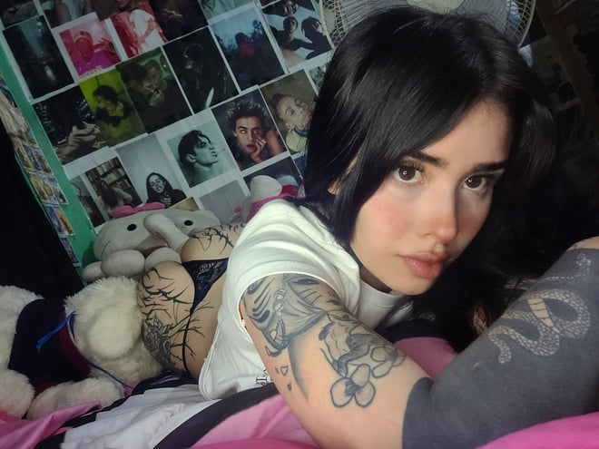 A young woman with dark hair and visible tattoos relaxing among plush toys and photo collages.