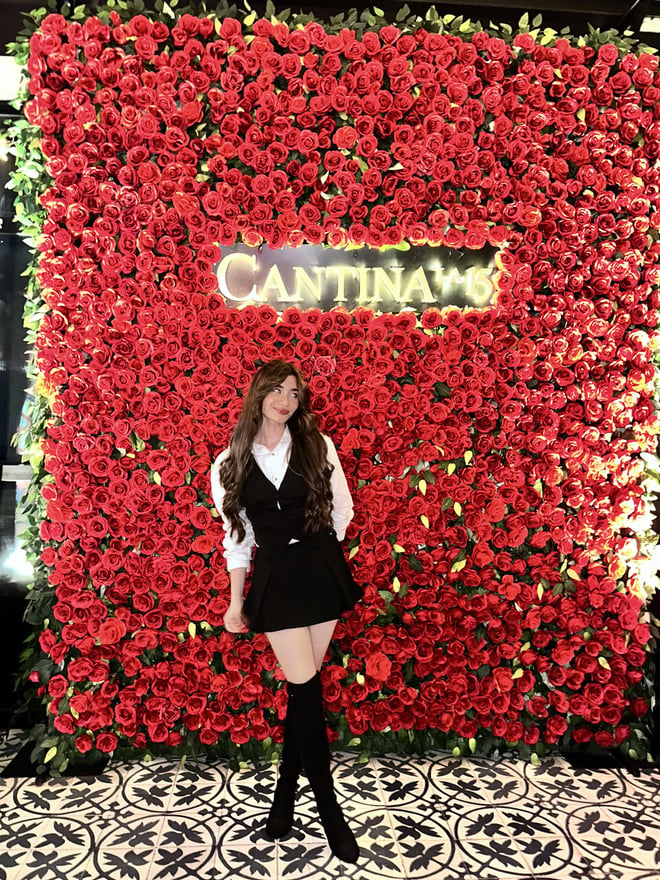 A young woman in a black outfit poses in front of a vibrant red rose wall with a restaurant sign.