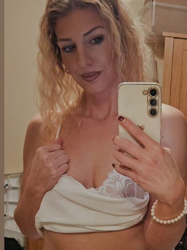 A blonde woman takes a selfie in a room, smiling with wavy hair, dressed in a light top and jewelry, standing near some boxes and furniture.