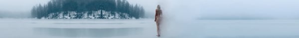 A silhouetted figure stands by a frozen lake surrounded by snowy trees in a foggy winter landscape.