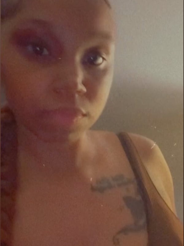 Close-up portrait of a woman with braids, makeup, and a tattoo visible on her chest, wearing a sleeveless top.