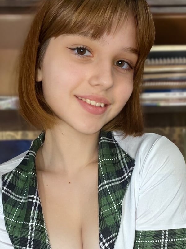Close-up of a young woman with short brown hair and smiling expression, dressed in a green plaid collared top against a bookshelf background.