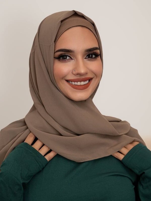A smiling woman wearing a beige hijab and green top, posing confidently against a plain background.