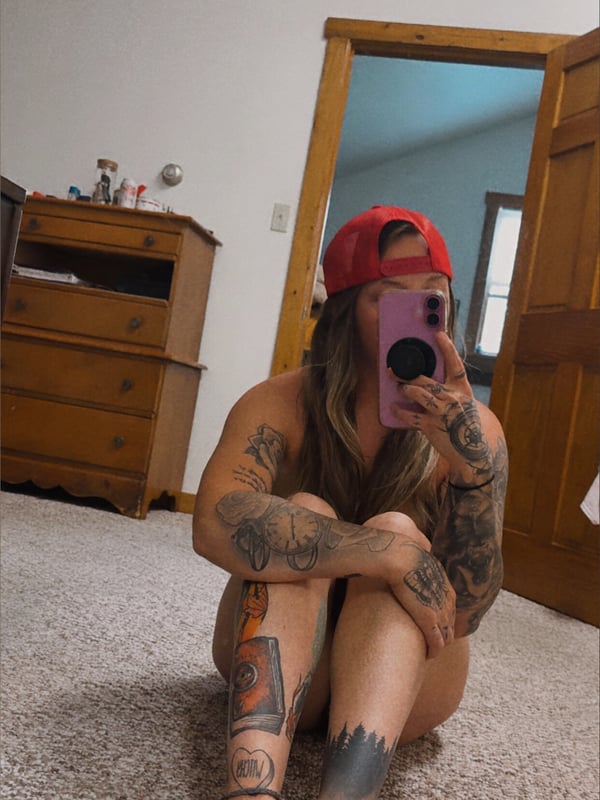 Woman with tattoos wearing a red cap, sitting on the floor in a bedroom with wooden furniture.