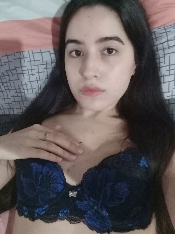 A young woman with long dark hair lying on a bed, wearing a lace top and looking at the camera.